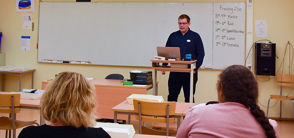 Lärare står framför tavlan och undervisar. I förgrunden sitter en elevassistent och en elev.